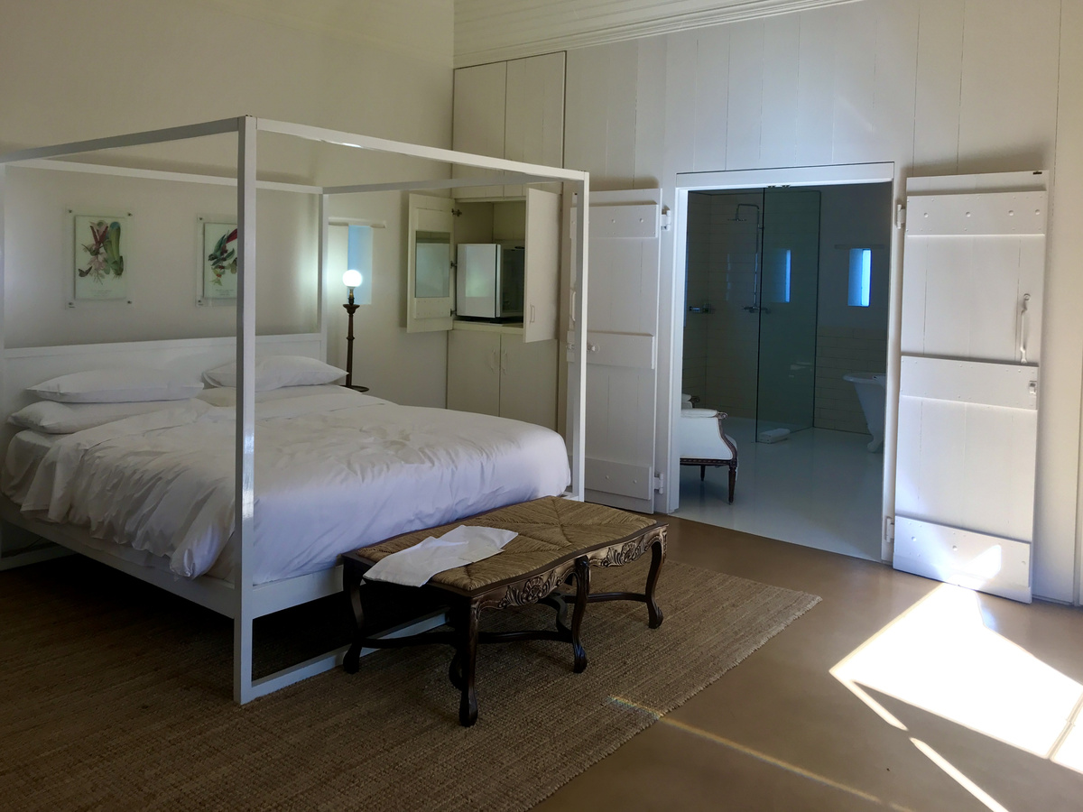 Wide view of a white four-poster bed, linen sofa and pale cupboards in a Babylonstoren cottage.