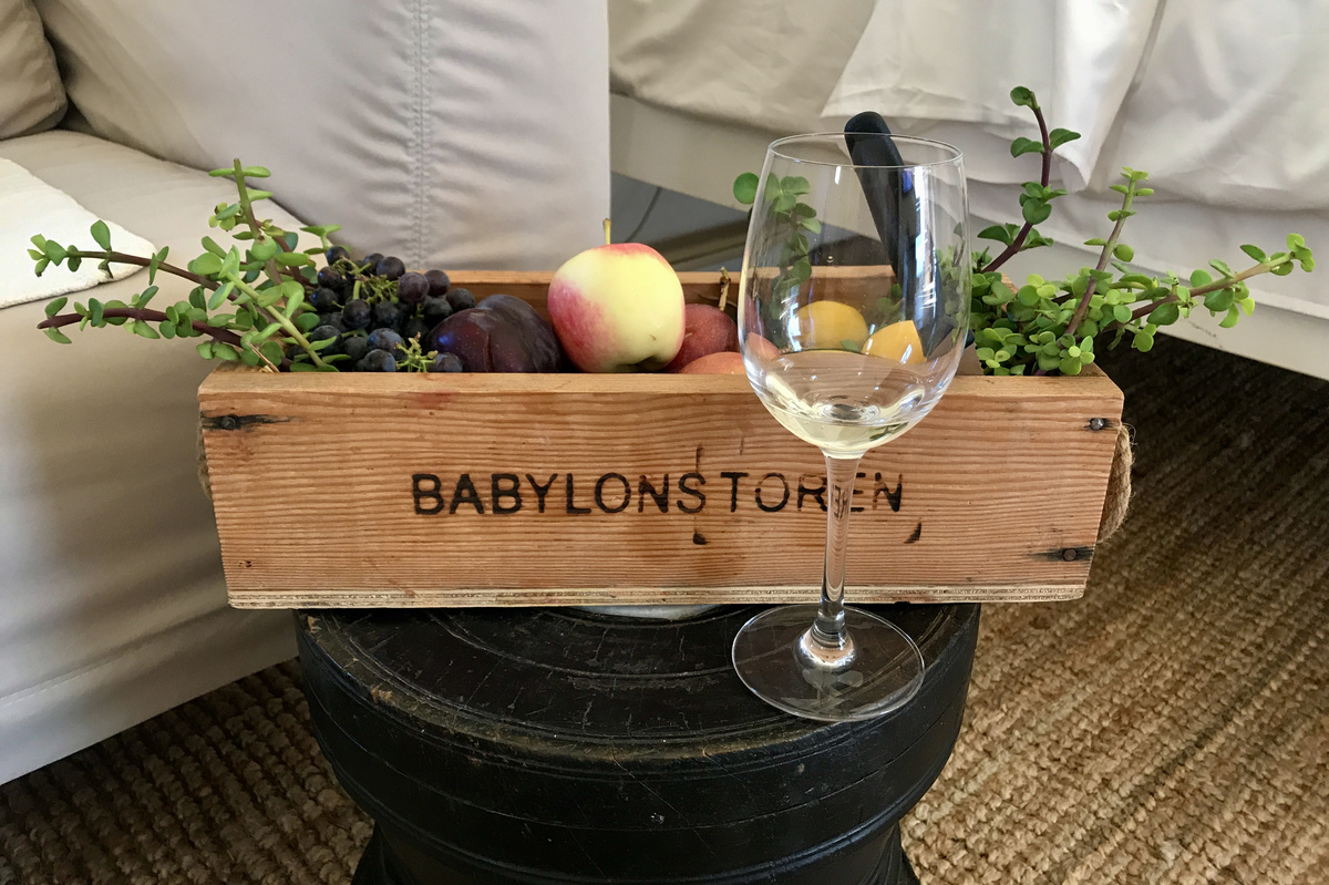 A wooden Babylonstoren crate holds grapes, plums, an apple and greenery beside a wine glass.