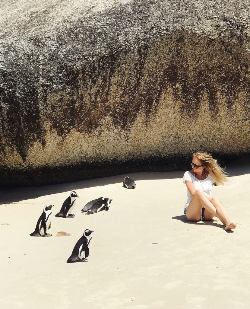 Olga sitting on pale sand beside five African penguins beneath a massive granite boulder at Boulders Beach.