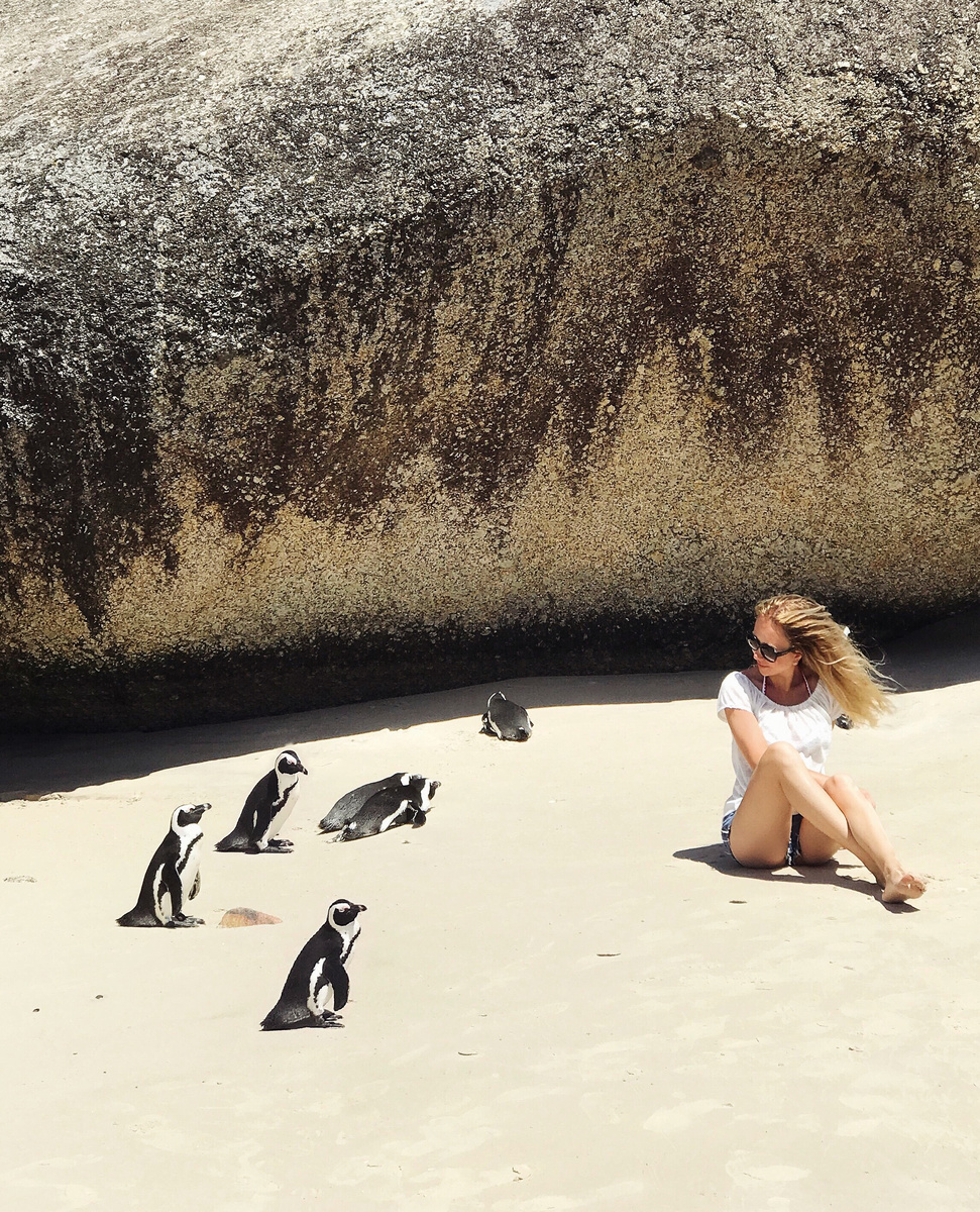 Olga sitting on pale sand beside five African penguins beneath a massive granite boulder at Boulders Beach.