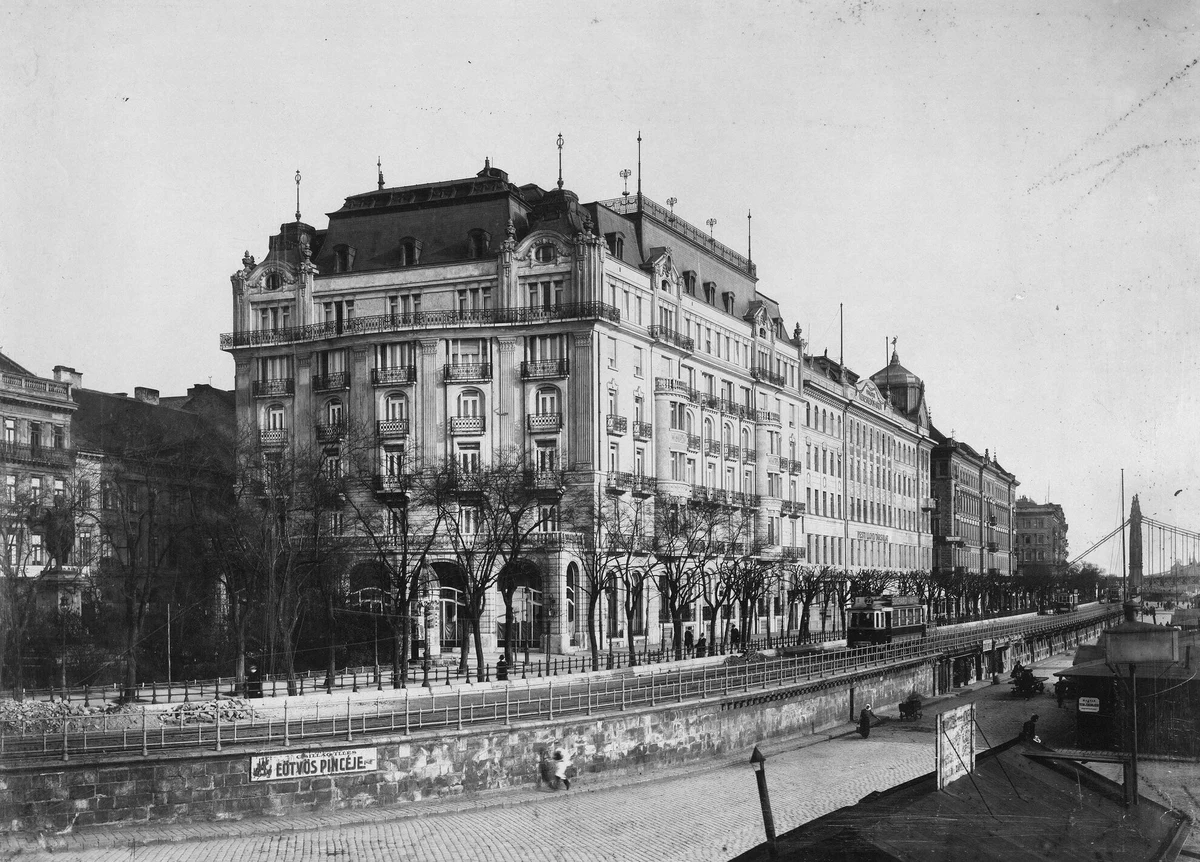 Black-and-white photograph of the Ritz Hotel Budapest facing the Danube in 1916, with pedestrians and early motorcars outside.