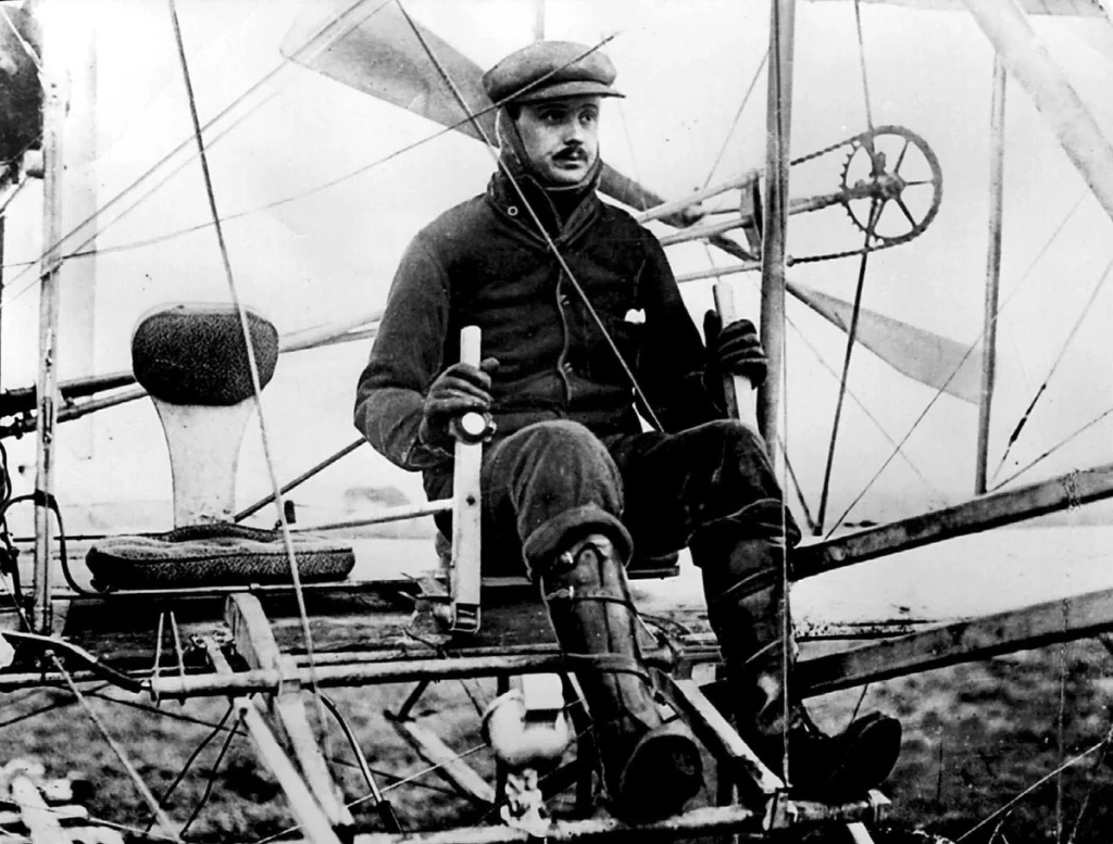 Charles Rolls sits at the controls of an early Wright-type biplane amid its exposed wires and wooden structure.