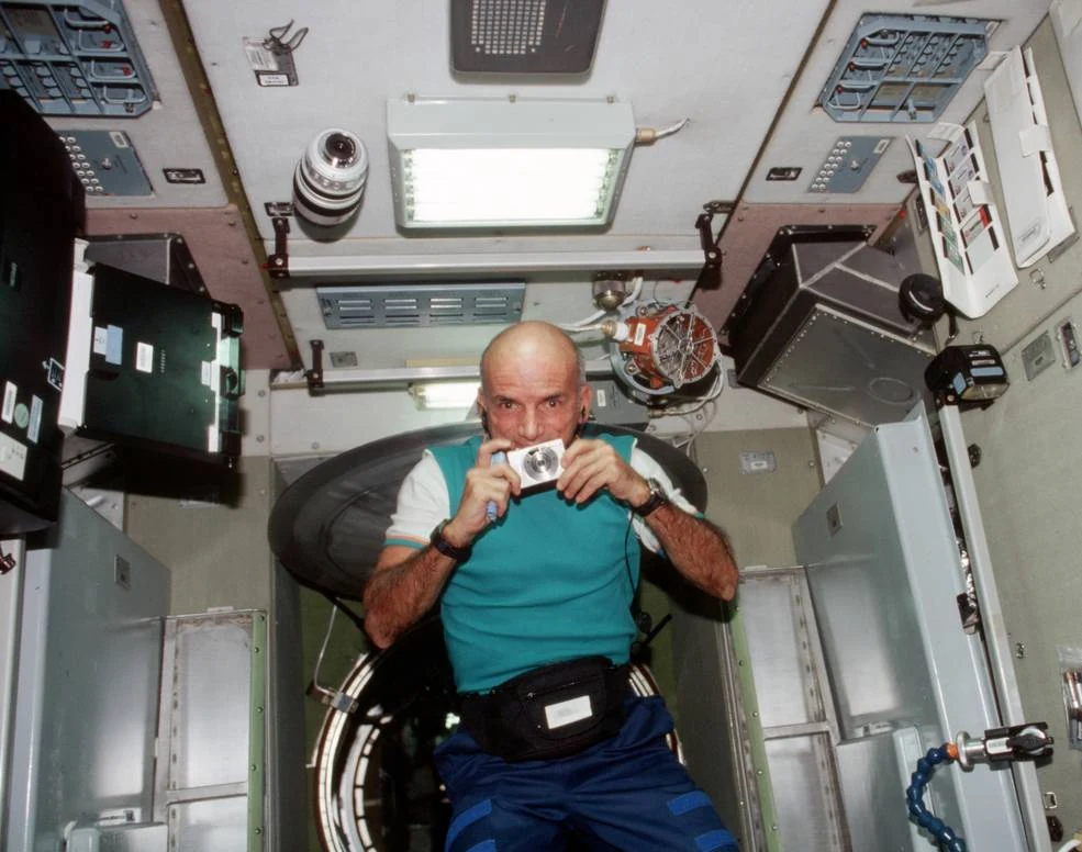 Dennis Tito floats inside the International Space Station and points a compact silver camera toward the viewer.