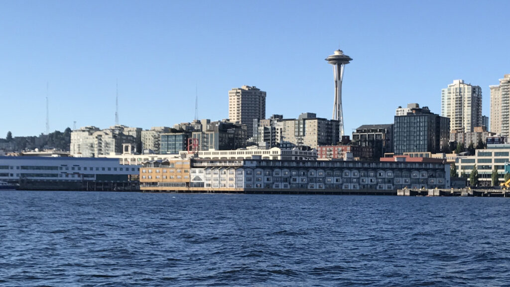 The Edgewater Hotel stretches across Pier 67 beneath its red neon sign and the Space Needle, seen from Elliott Bay.