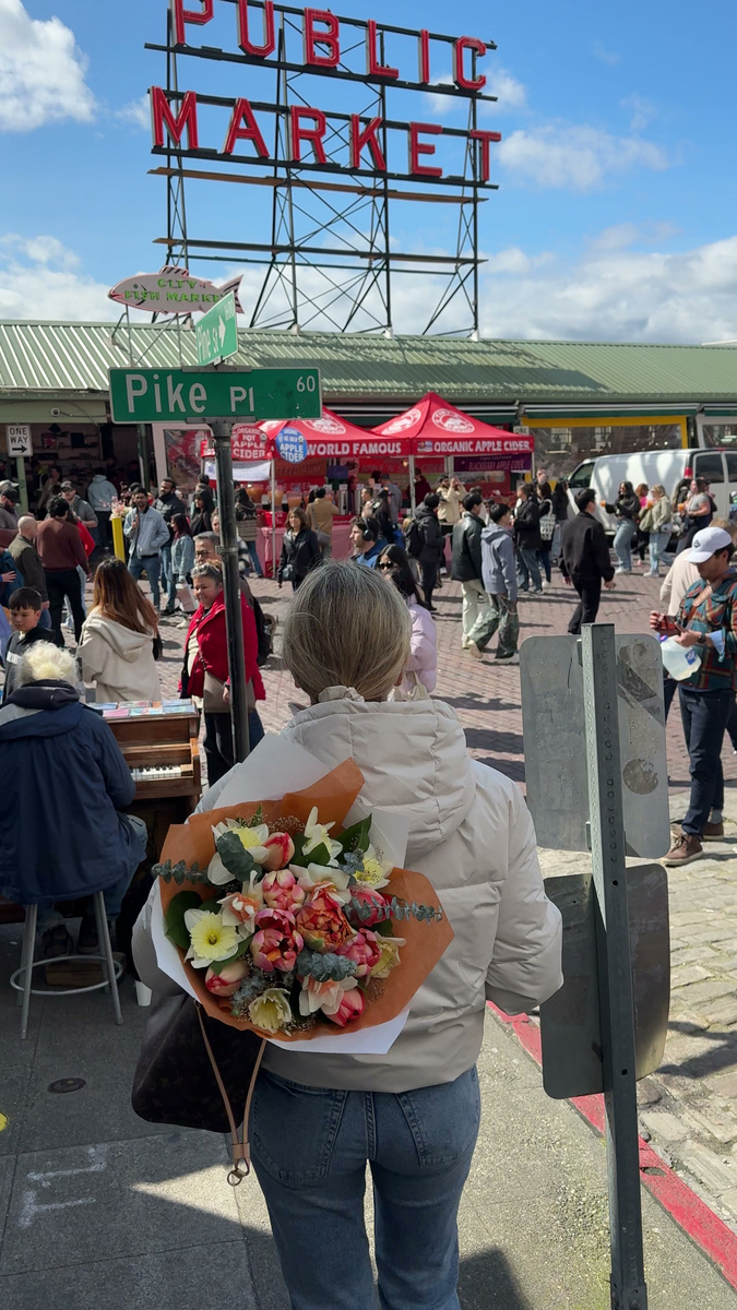 Olga walks beneath the red Public Market sign at Pike Place Market in Seattle, carrying a bright bouquet.