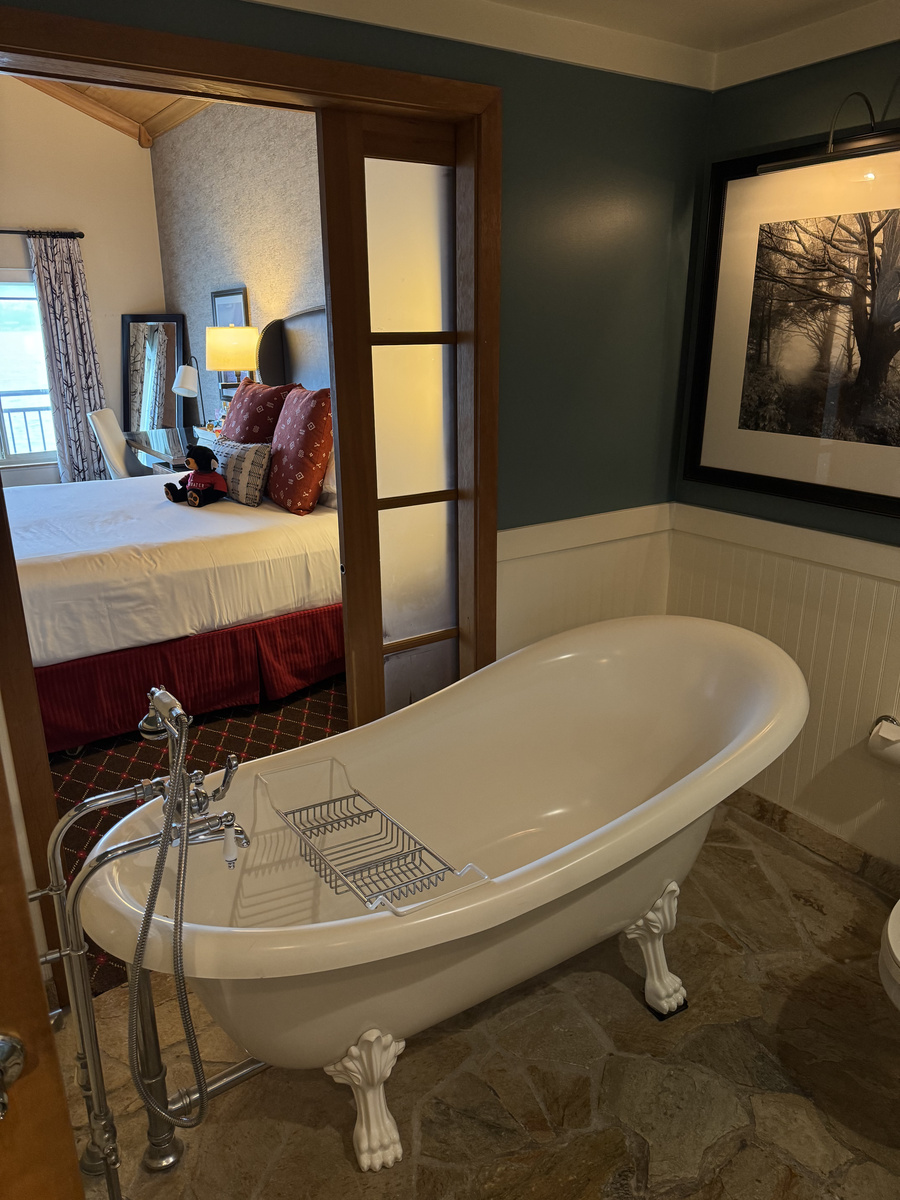 A white claw-foot bathtub in an Edgewater Hotel guest room, with a sliding wooden screen opening toward the bed and waterfront window.