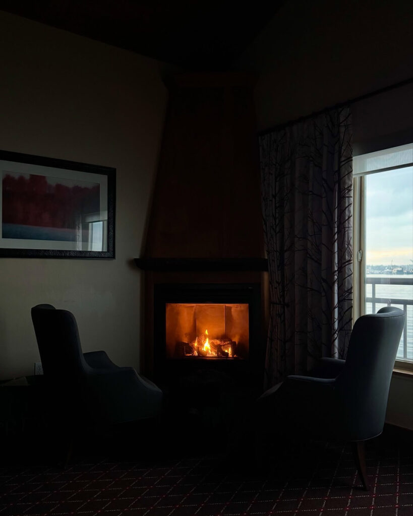 A lit gas fireplace and two dark chairs stand beside an over-water window at The Edgewater Hotel.