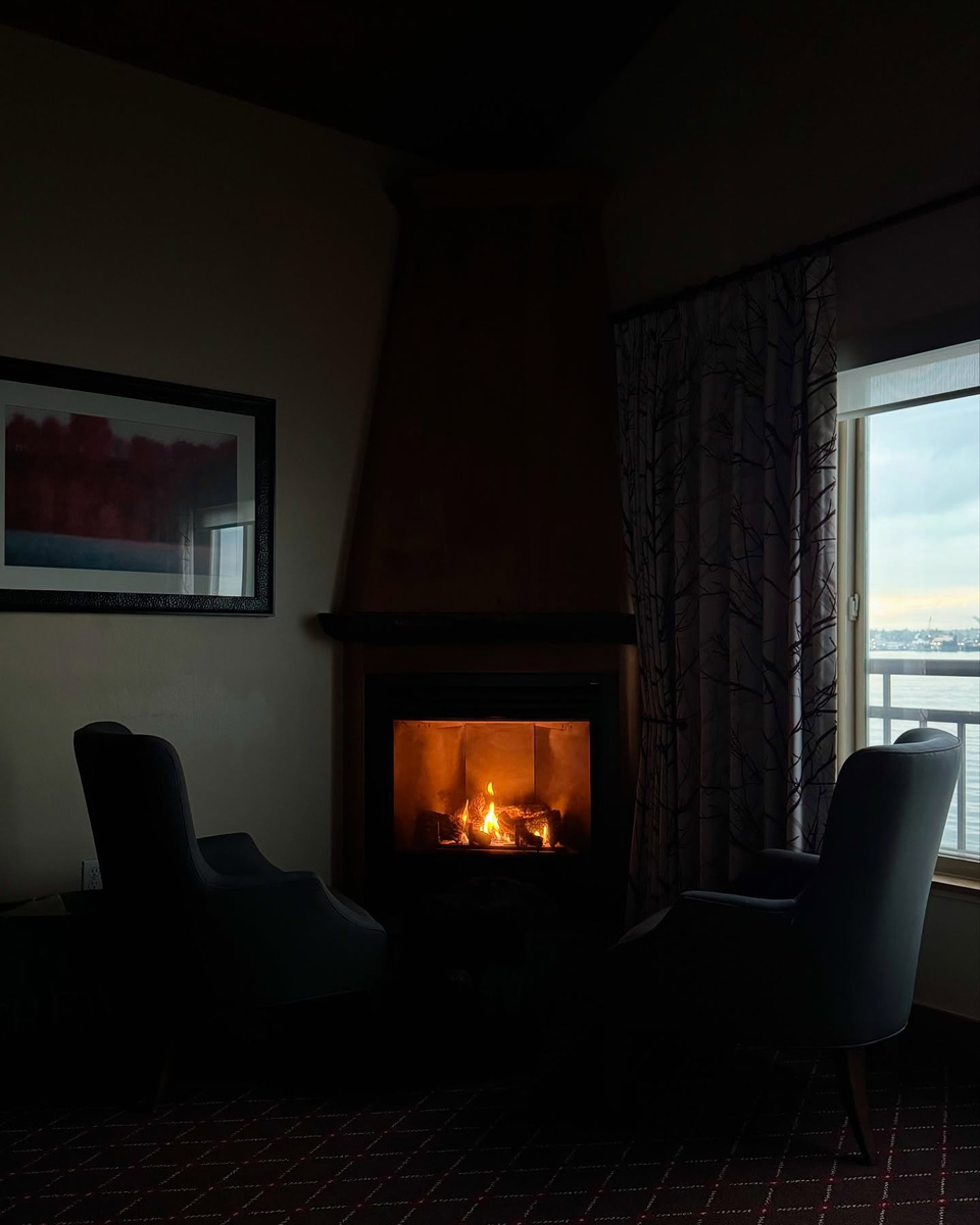 A lit gas fireplace and two dark chairs stand beside an over-water window at The Edgewater Hotel.