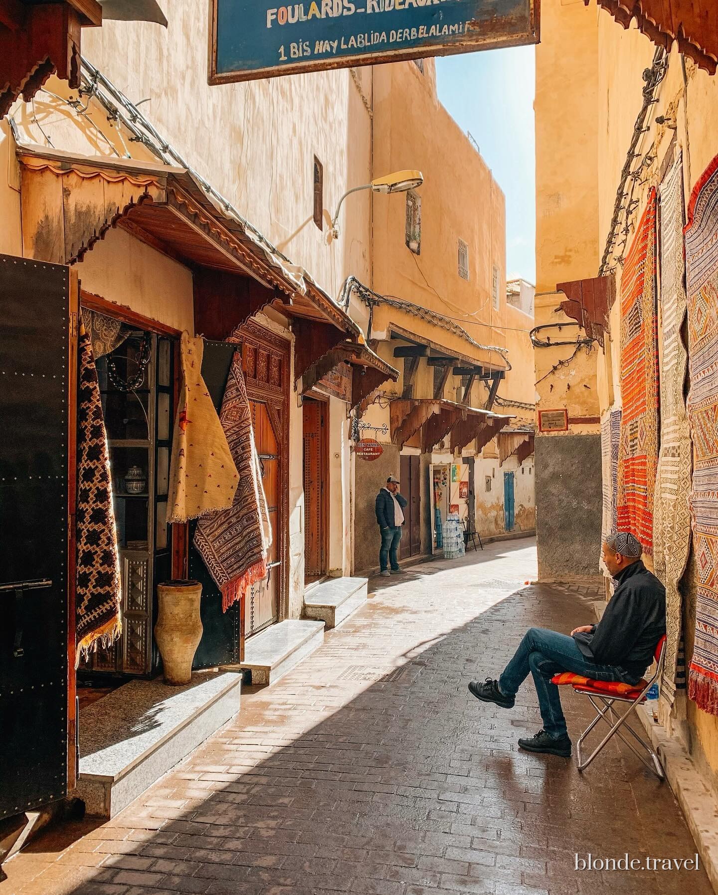 A man sits beside carpet shops in a narrow sunlit lane of the Fez medina.