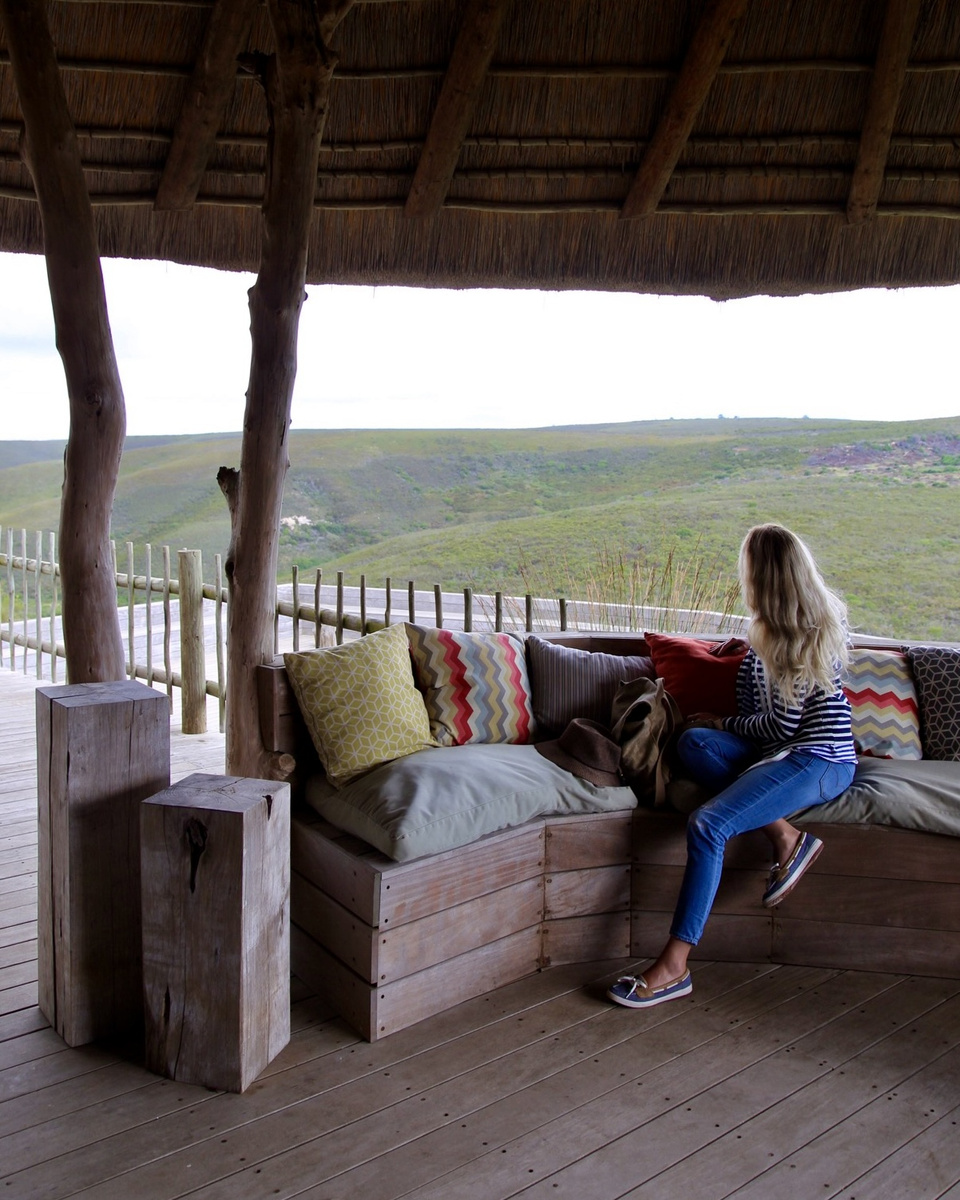 Olga sitting on a cushioned wooden sofa on a thatched veranda overlooking green hills