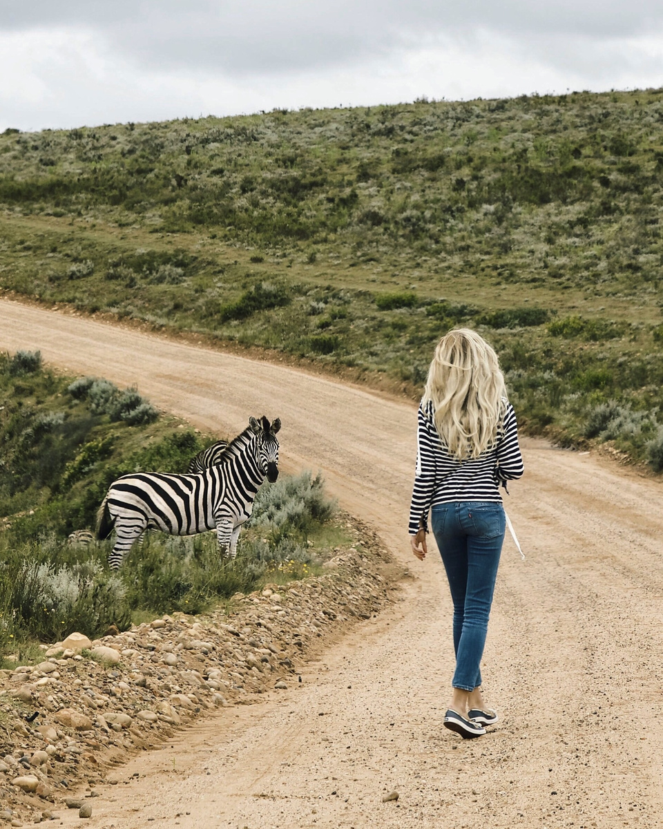 Olga walking along a dirt road with a zebra standing in the vegetation beside her