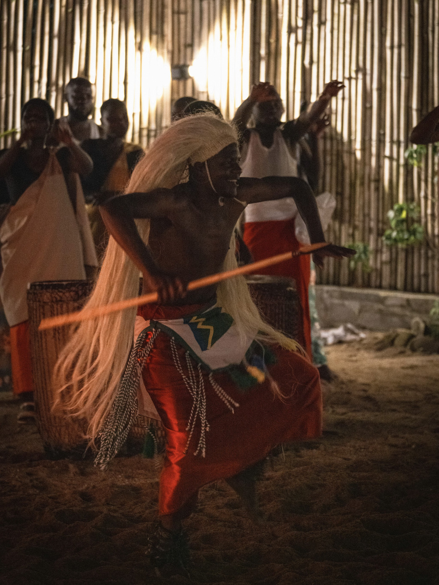 A Gorilla Guardians dancer in layered textiles and raffia moves before a row of musicians.