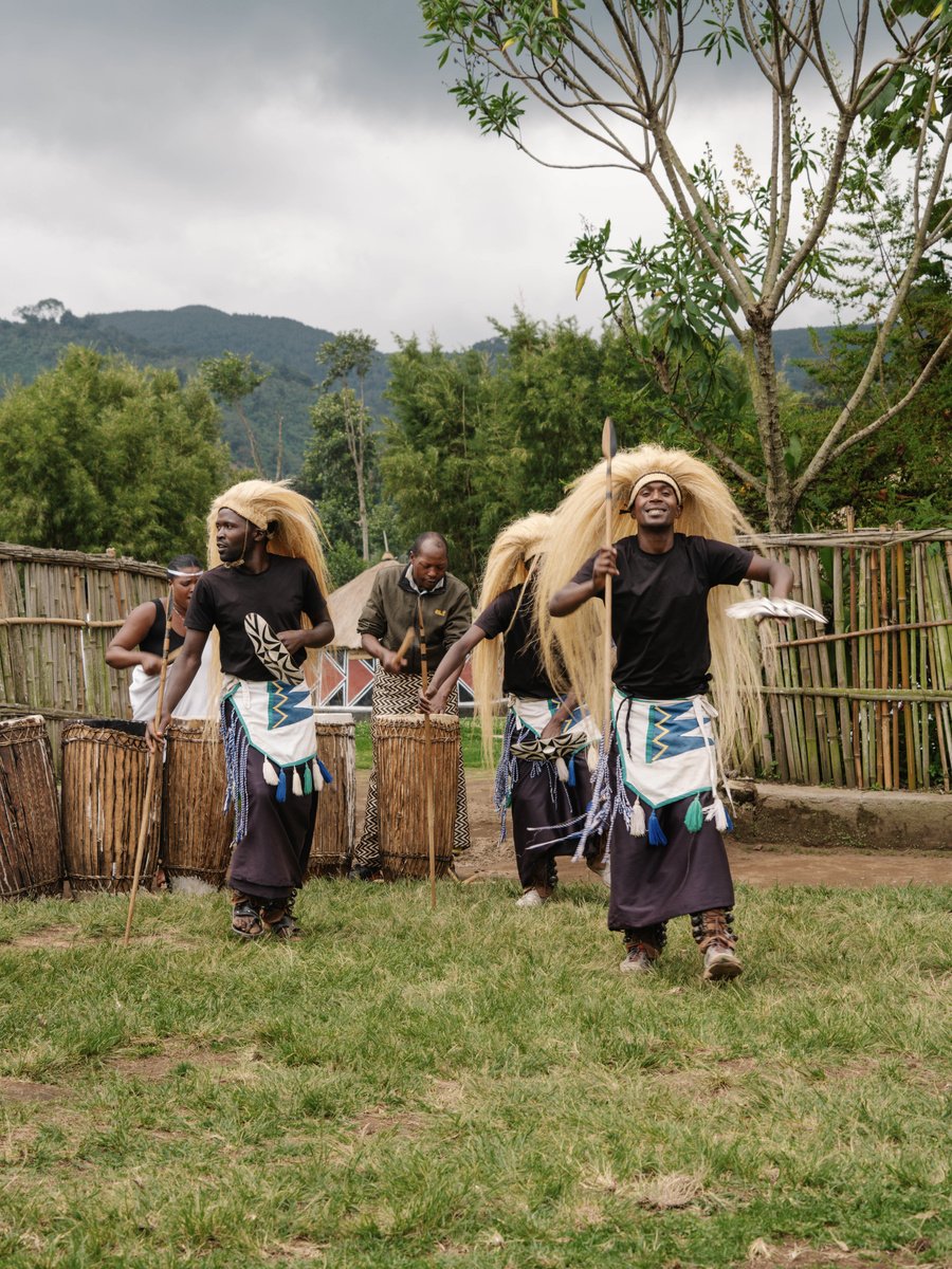 Gorilla Guardians performers dance outdoors in traditional dress with woven fencing behind them.