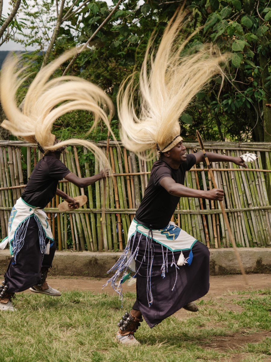 Two Gorilla Guardians dancers swing long pale headdresses as they move across a grass performance area.