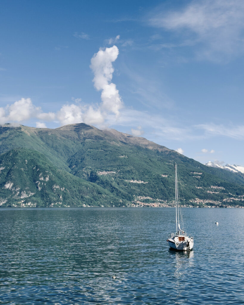 A white sailboat rests on Lake Como below green mountains and a tall summer cloud.
