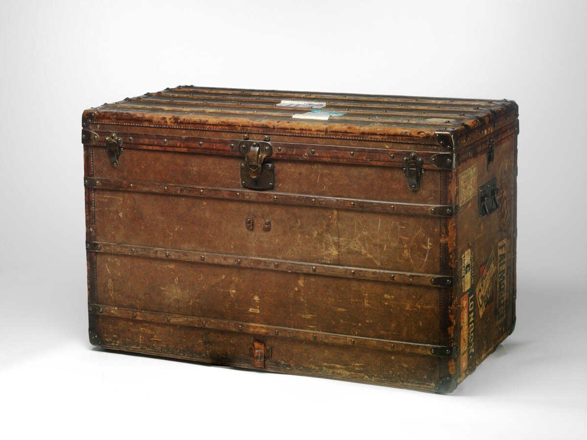 Large brown Louis Vuitton Malle Haute trunk with a flat slatted top, metal fittings, scuffed surfaces and ocean-liner labels.