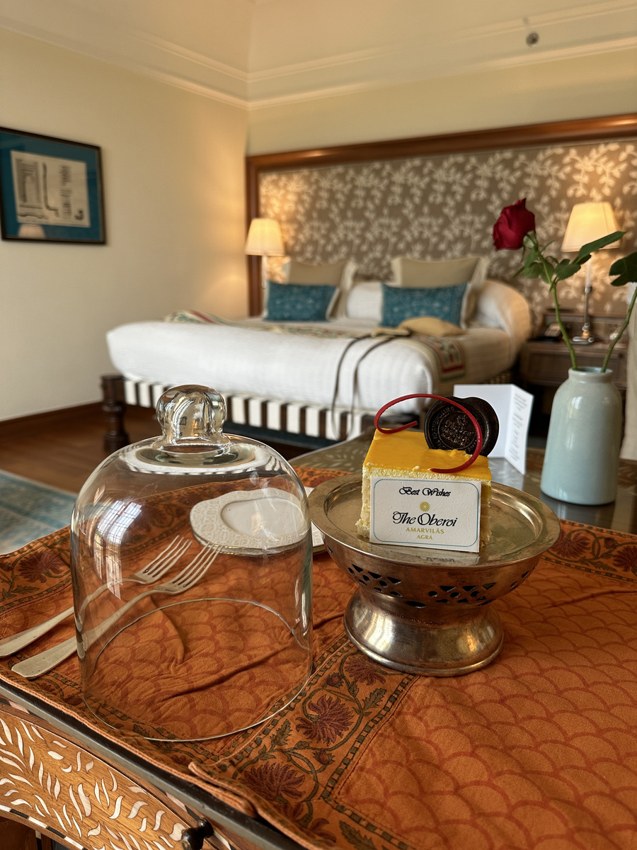 A small Oberoi Amarvilas birthday cake sits on a table with red roses and the bed behind.