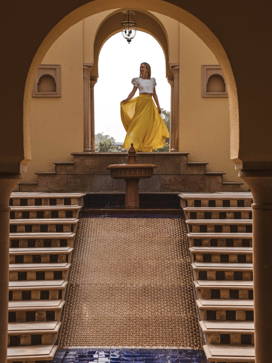 Olga in a yellow skirt stands above a stepped water cascade beneath an arch at The Oberoi Amarvilas.