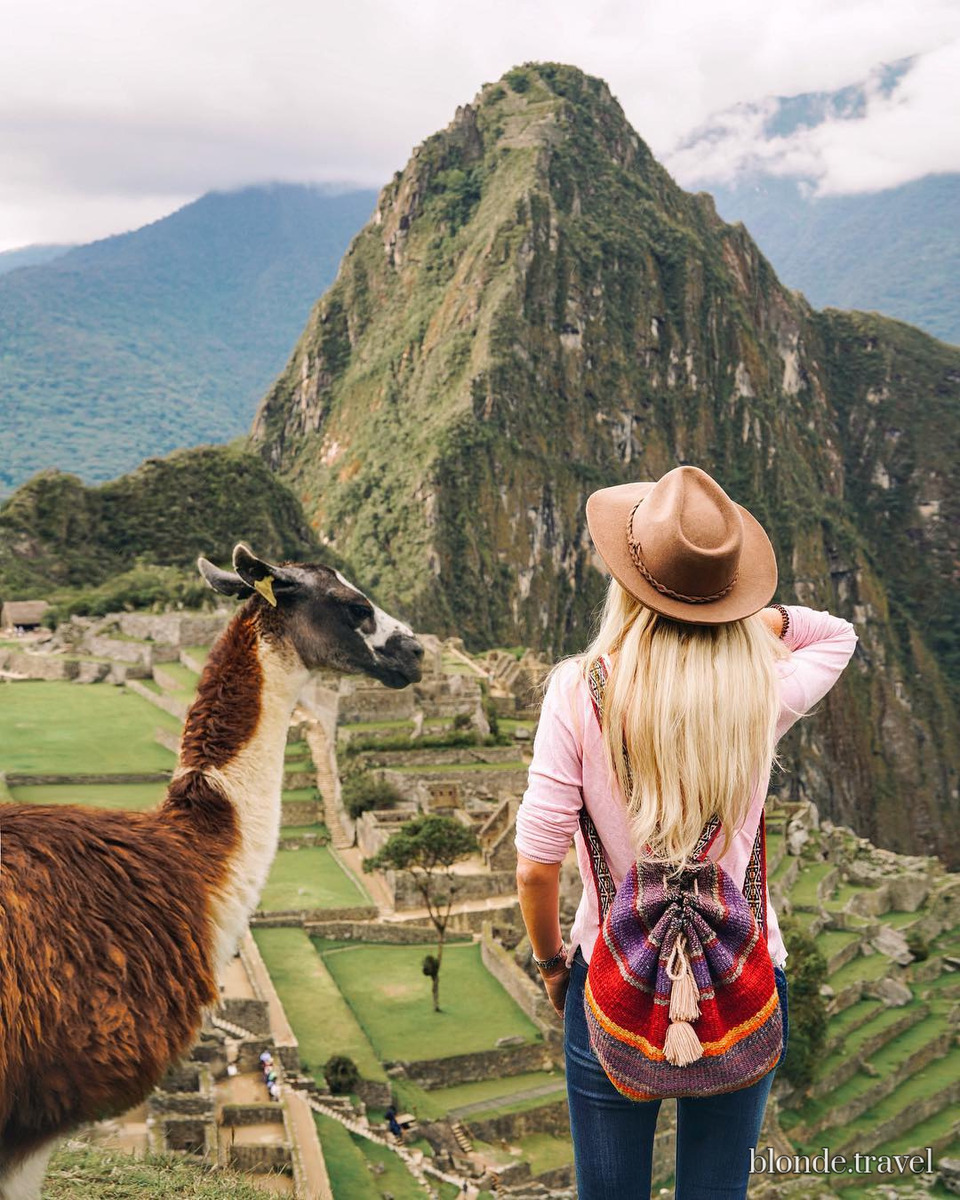 Olga stands beside a llama overlooking the stone terraces and green peaks of Machu Picchu.