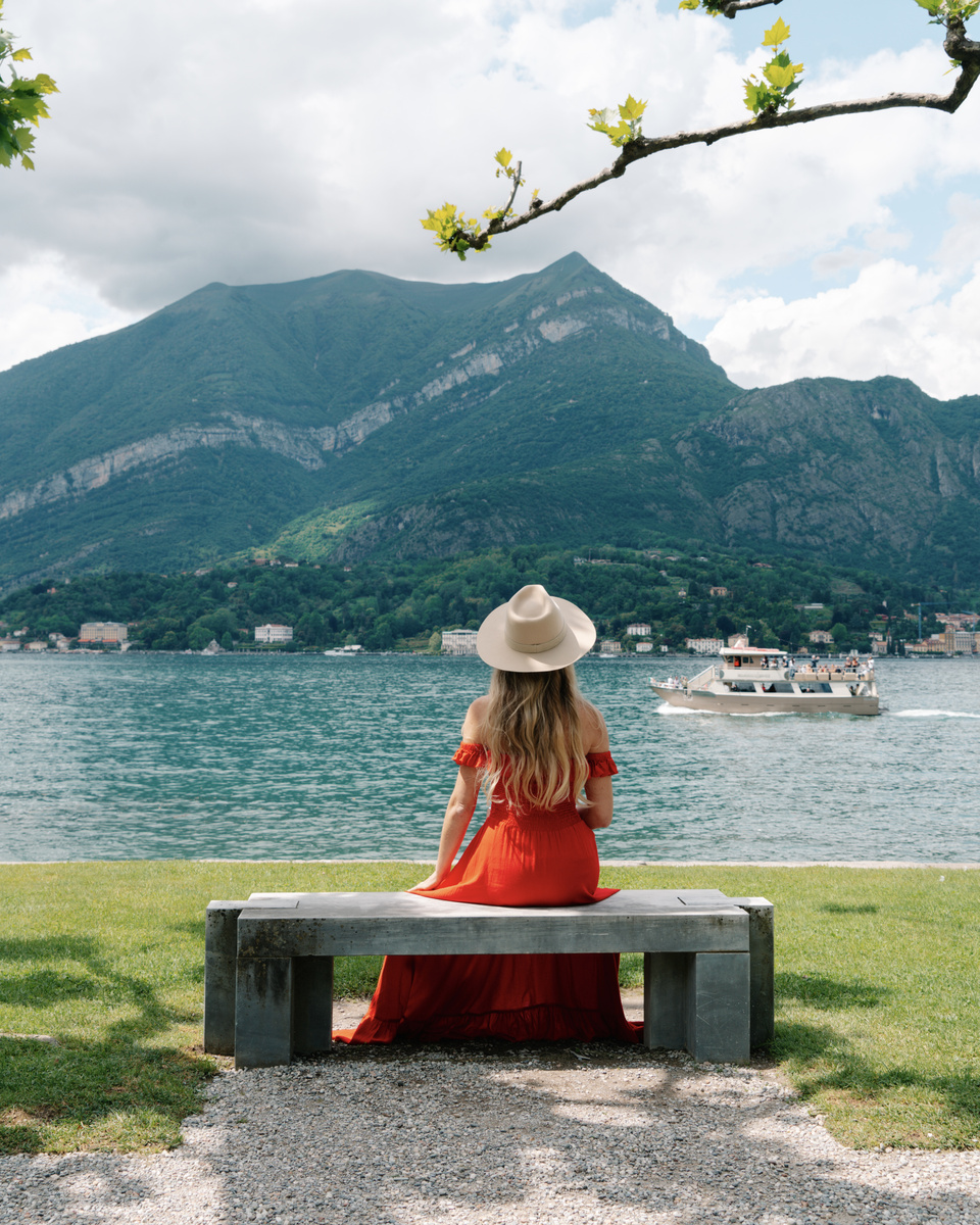 Olga, wearing a red dress and straw hat, sits on a bench in the gardens of Villa Melzi as a passenger boat crosses Lake Como beneath the mountains.