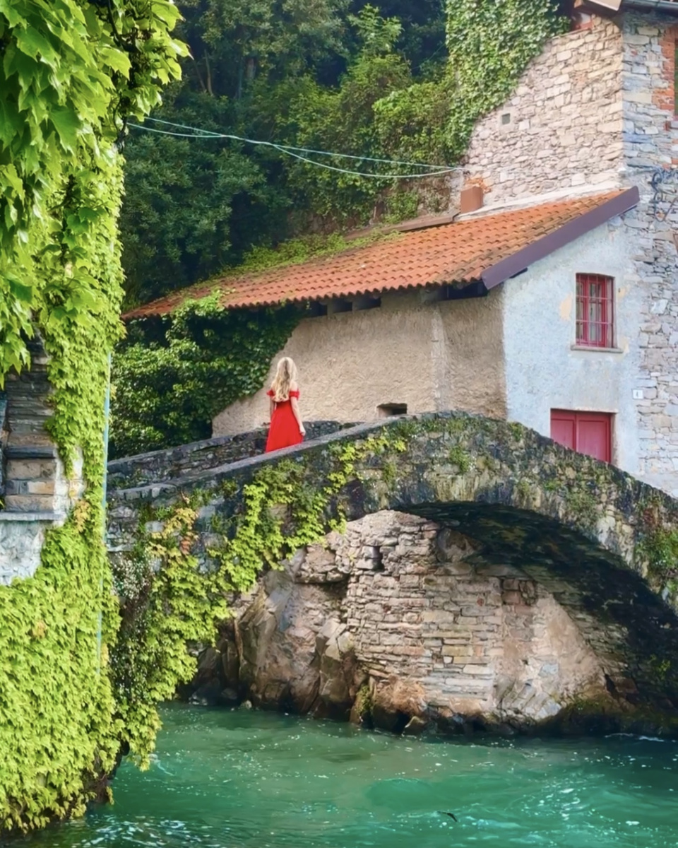 Olga, wearing a red dress, walks across Ponte della Civera in Nesso, surrounded by ivy and trees.