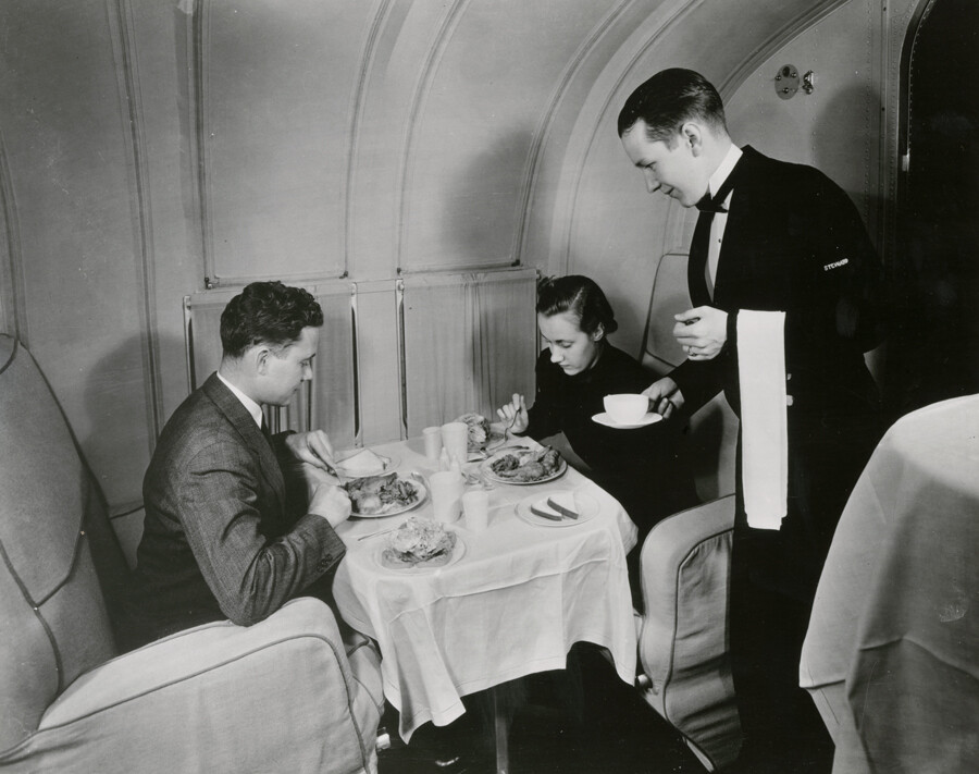 A steward serves dinner to two formally dressed passengers seated at a linen-covered table aboard a Pan American Martin M-130 flying boat.