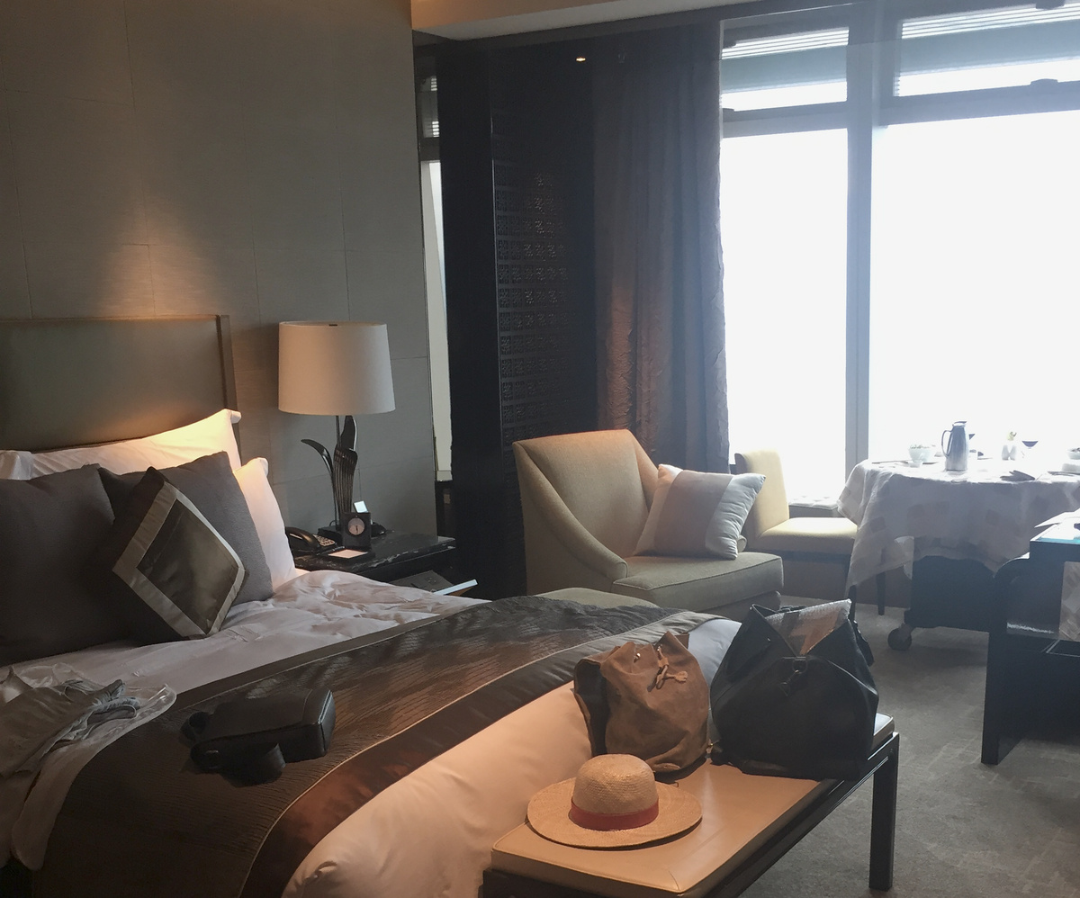 A Ritz-Carlton Hong Kong room with a bed, window seat and breakfast table beside the harbour-facing windows