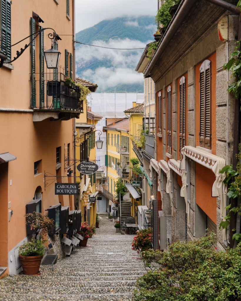 Salita Serbelloni descends between ochre and terracotta buildings toward misty mountains in Bellagio.