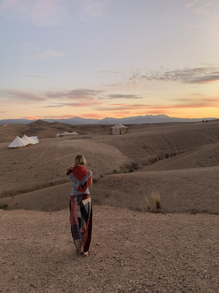 Olga walking above Scarabeo Camp at sunset, with canvas tents and the Atlas Mountains ahead