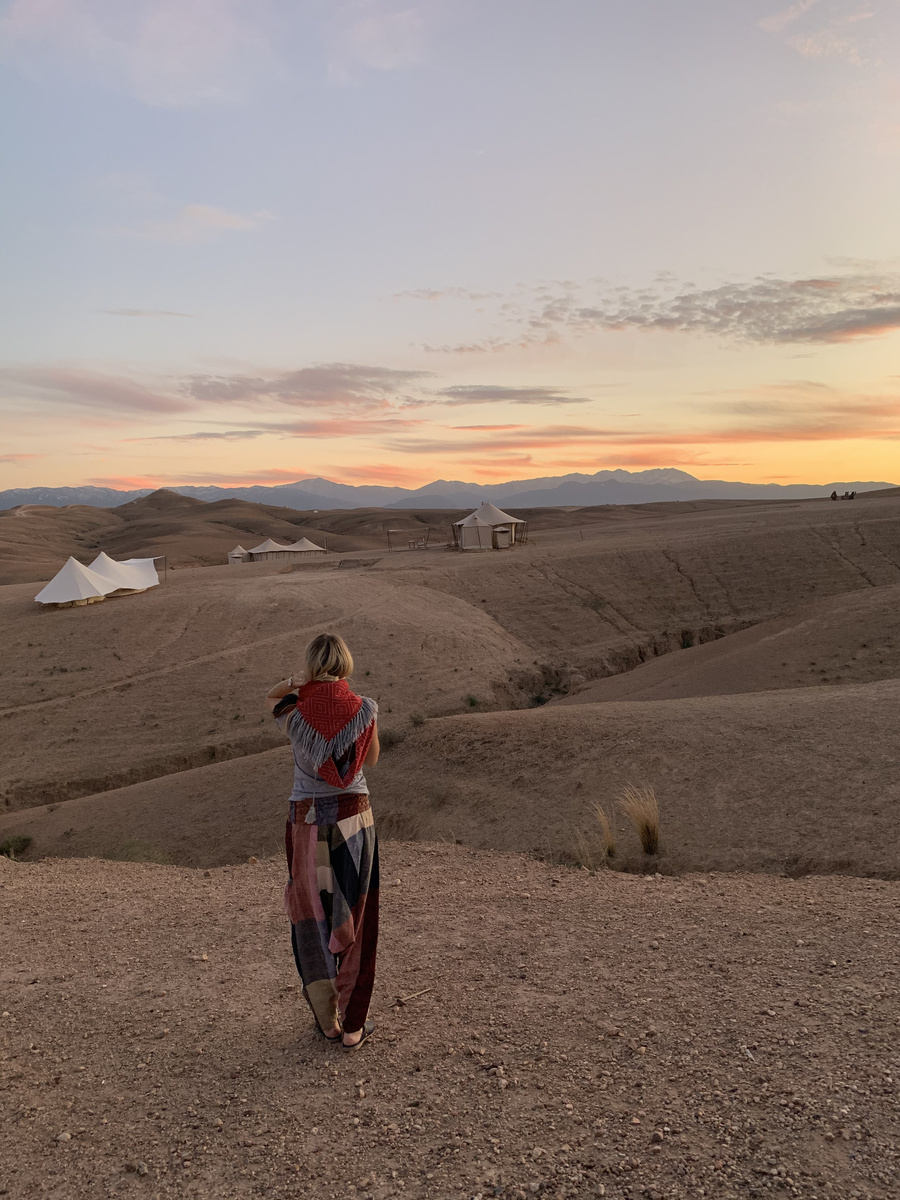 Olga walking above Scarabeo Camp at sunset, with canvas tents and the Atlas Mountains ahead