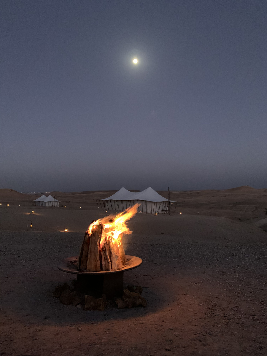 Wood fire beneath the moon at Scarabeo Camp, with illuminated tents across the dark Agafay landscape