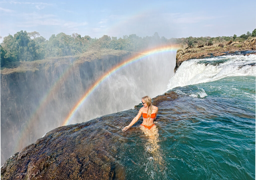 Olga stands in a rock pool at the edge of Victoria Falls with a rainbow across the gorge.