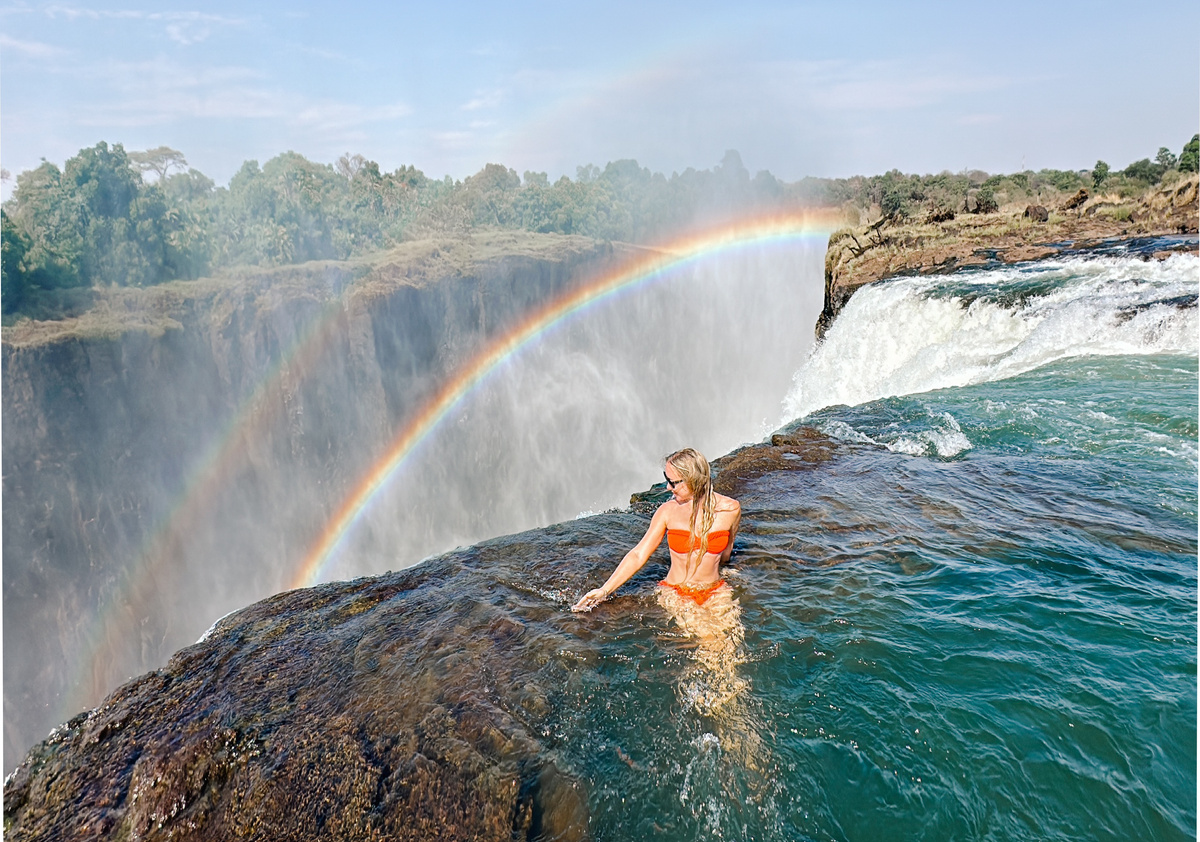 Olga stands in a rock pool at the edge of Victoria Falls with a rainbow across the gorge.