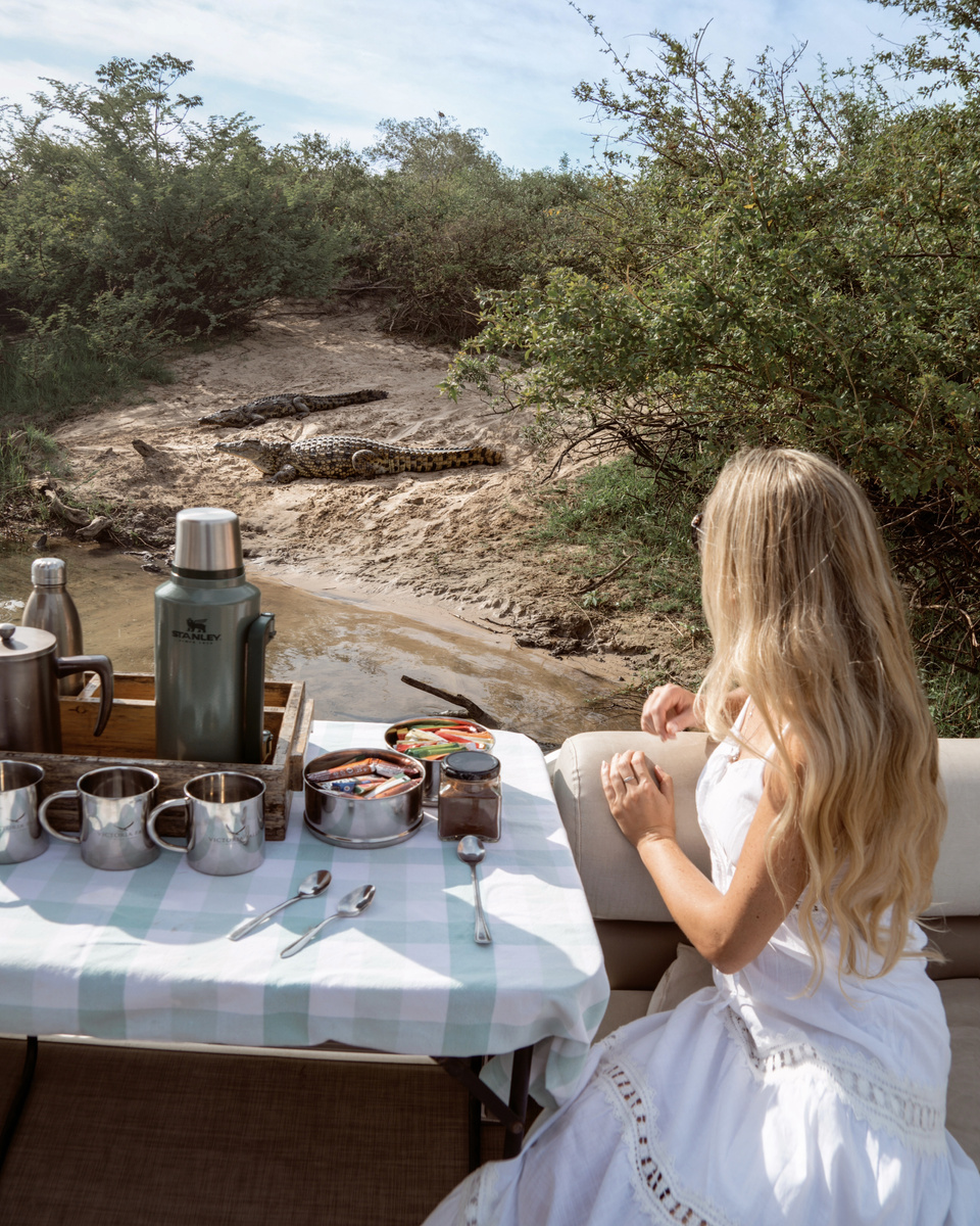 Olga sits at an outdoor breakfast table with coffee and metal serving pots beside the bush near Victoria Falls River Lodge.