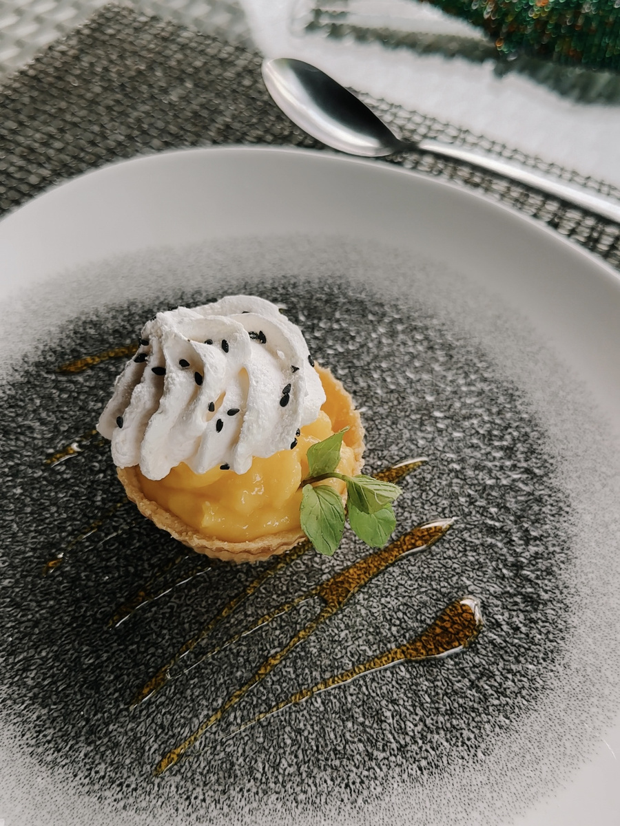 A small yellow tart topped with white cream, black seeds and mint is served on a dark plate at Victoria Falls River Lodge.