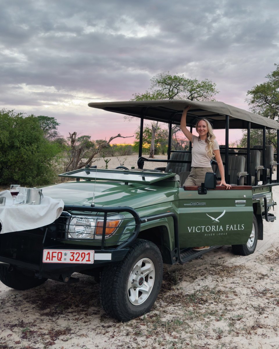 Olga stands beside an open Victoria Falls River Lodge game-drive vehicle at sunset.