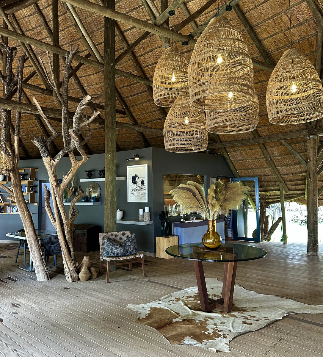 The open main lounge at Victoria Falls River Lodge has a high thatched roof, timber supports, woven pendant lights and locally inspired art.