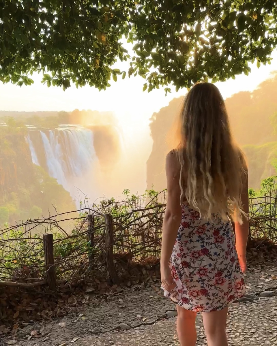 Olga stands beneath a tree as sunrise lights the mist and falling water at Victoria Falls.