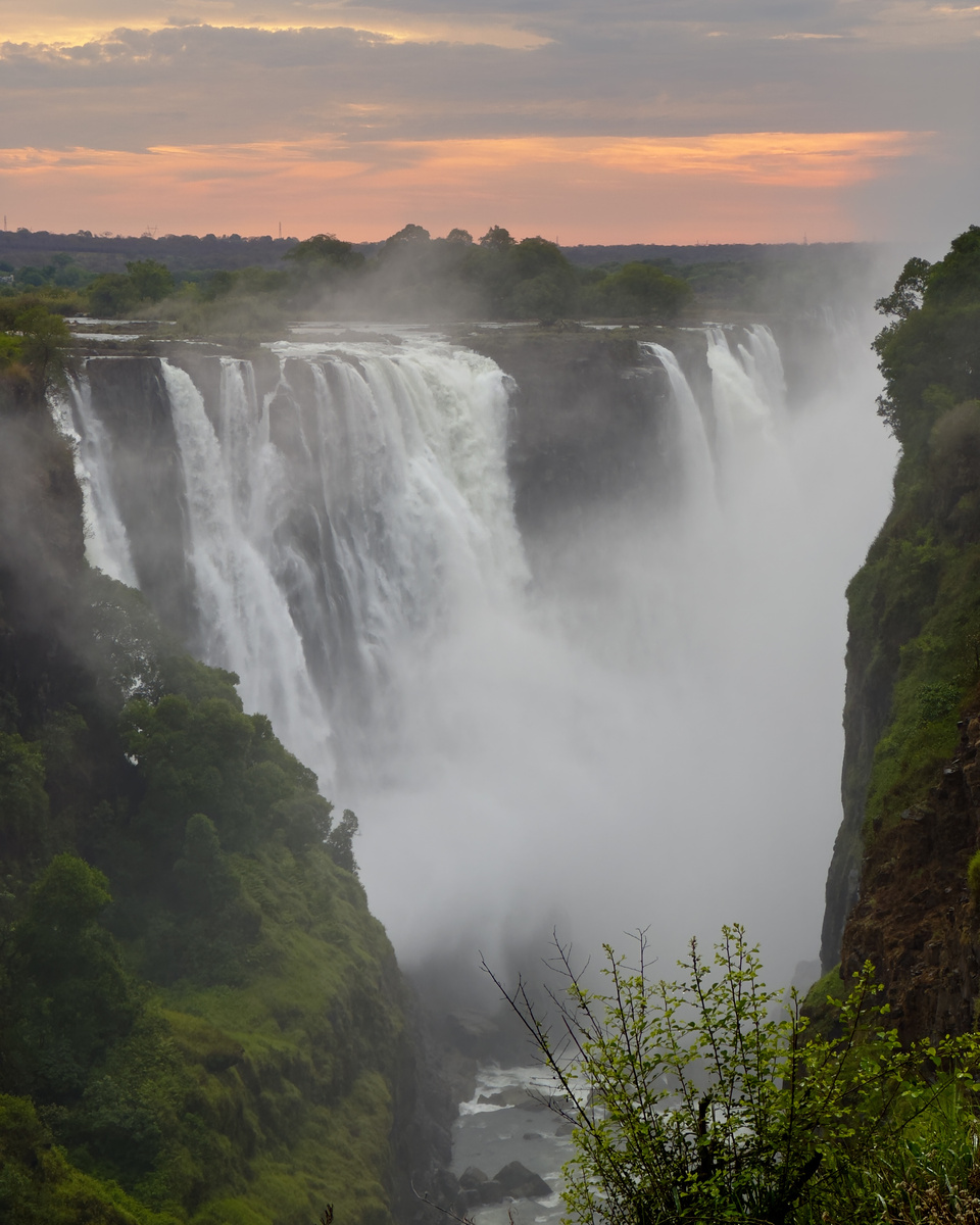Victoria Falls drops into its gorge beneath a pale orange sunrise and drifting mist.