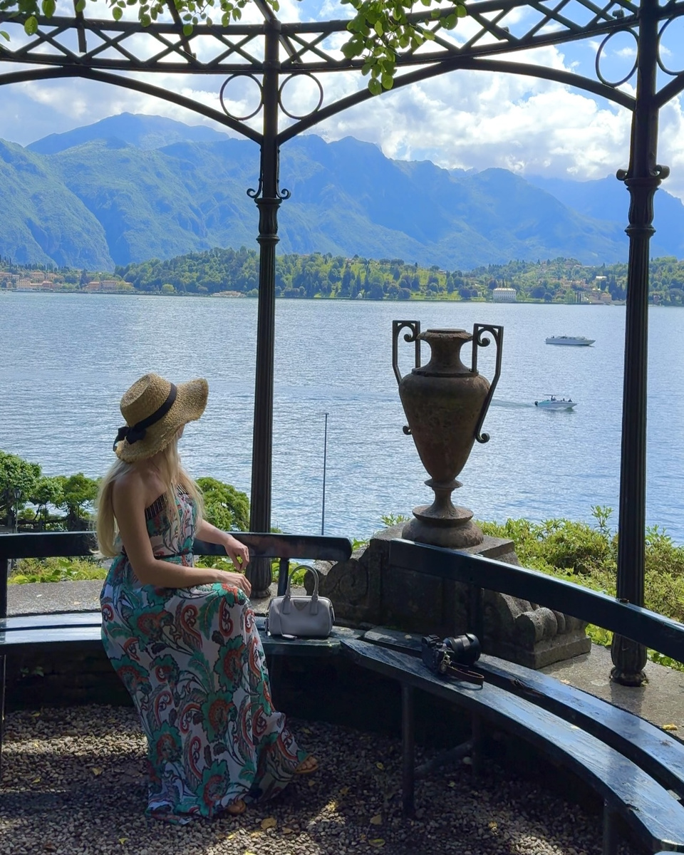 Olga, wearing a patterned dress and straw hat, looks across Lake Como from a garden pavilion at Villa Carlotta.