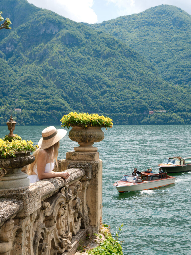 Olga, wearing a white dress and straw hat, looks across Lake Como from a carved stone terrace as two wooden taxi boats pass below.
