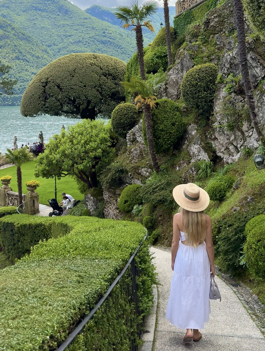 Olga, wearing a white dress and straw hat, walks through the clipped terraced gardens of Villa del Balbianello with Lake Como and mountains beyond.