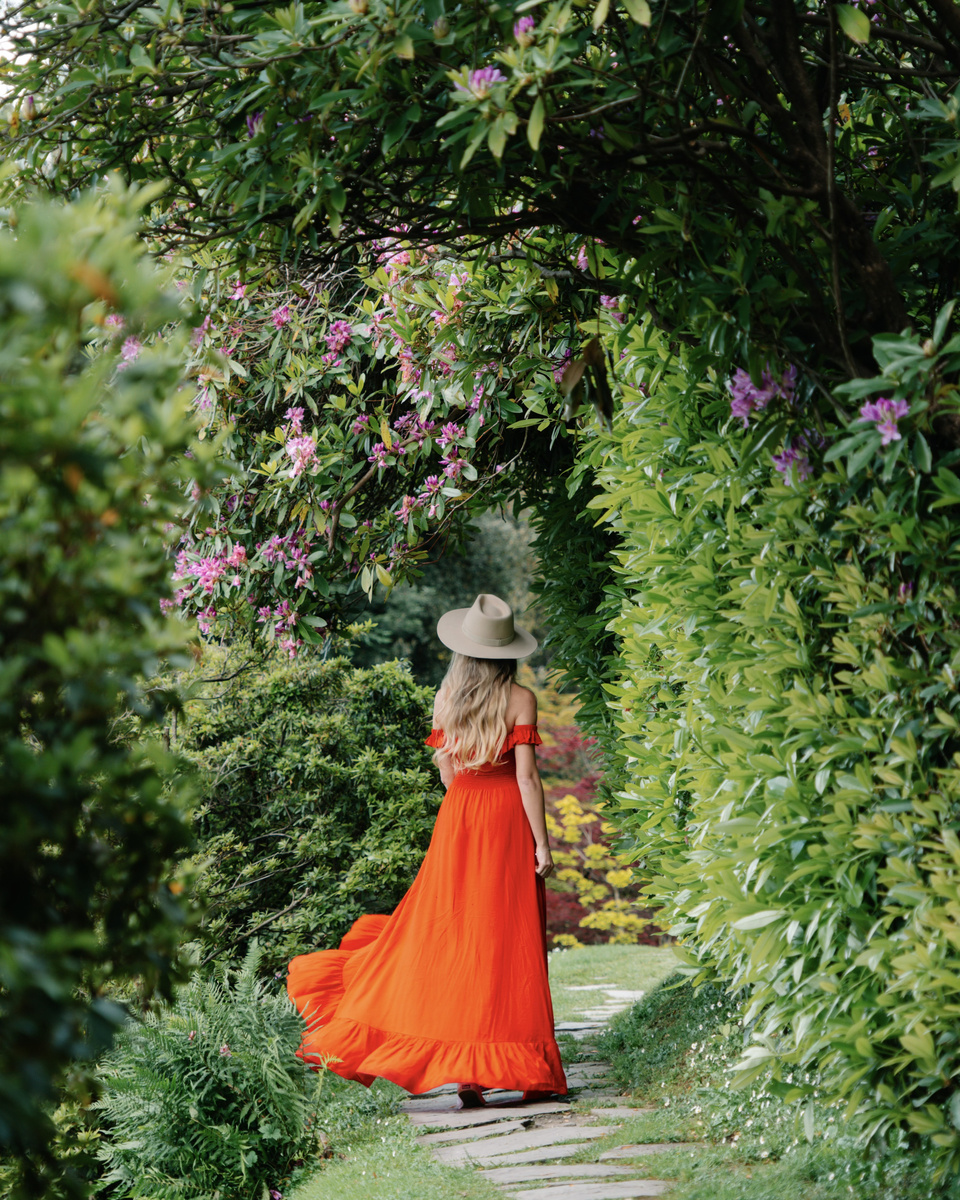 Olga, wearing a red dress and straw hat, walks beneath flowering rhododendrons in the gardens of Villa Melzi.