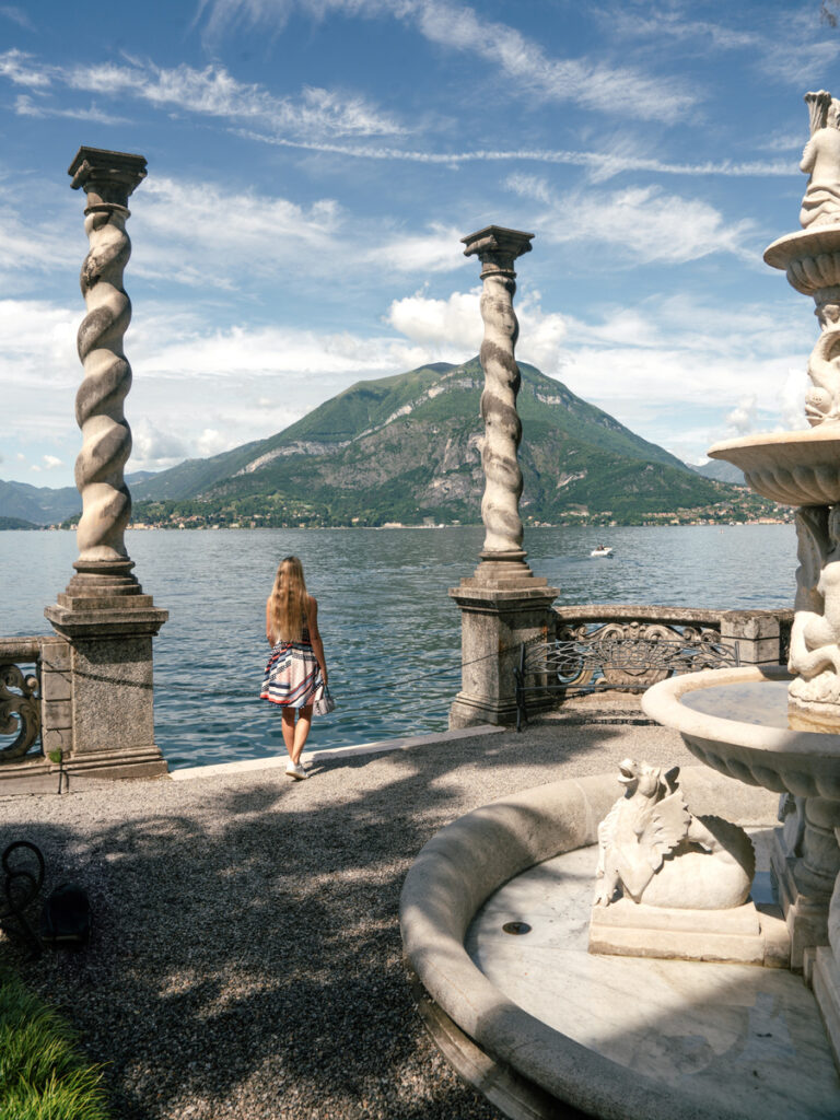 Olga stands between twisted stone columns beside a sculpted fountain overlooking Lake Como in the gardens of Villa Monastero.