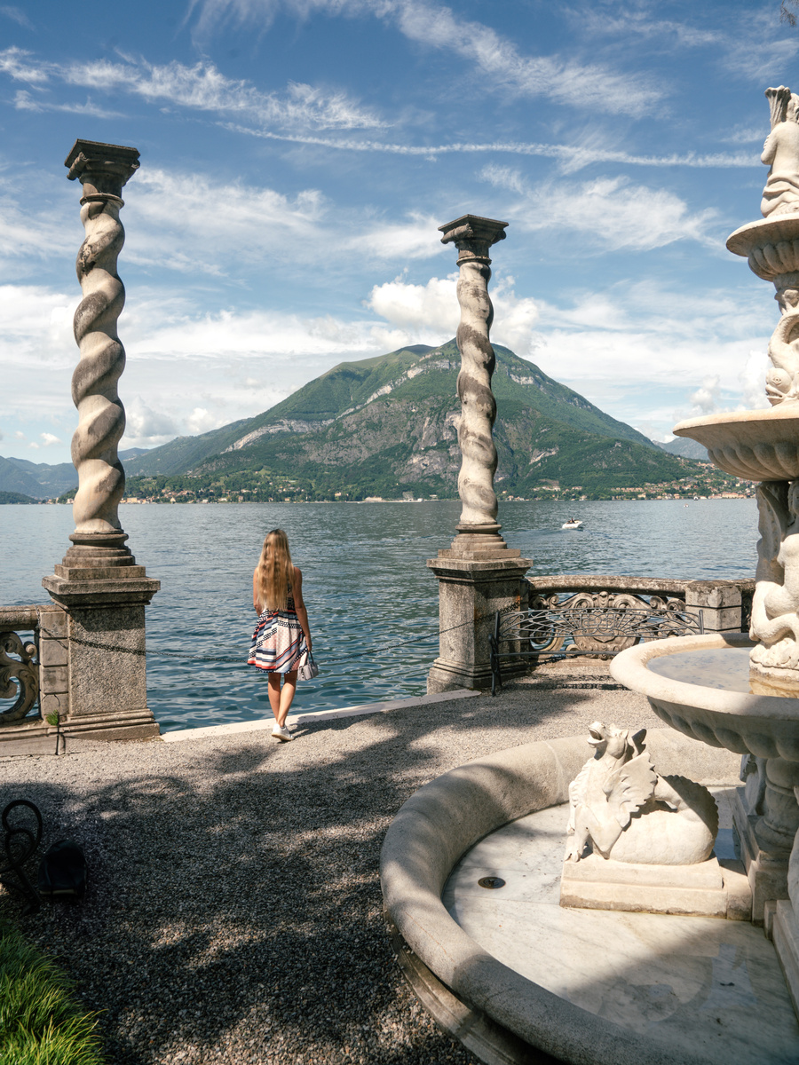 Olga stands between twisted stone columns beside a sculpted fountain overlooking Lake Como in the gardens of Villa Monastero.