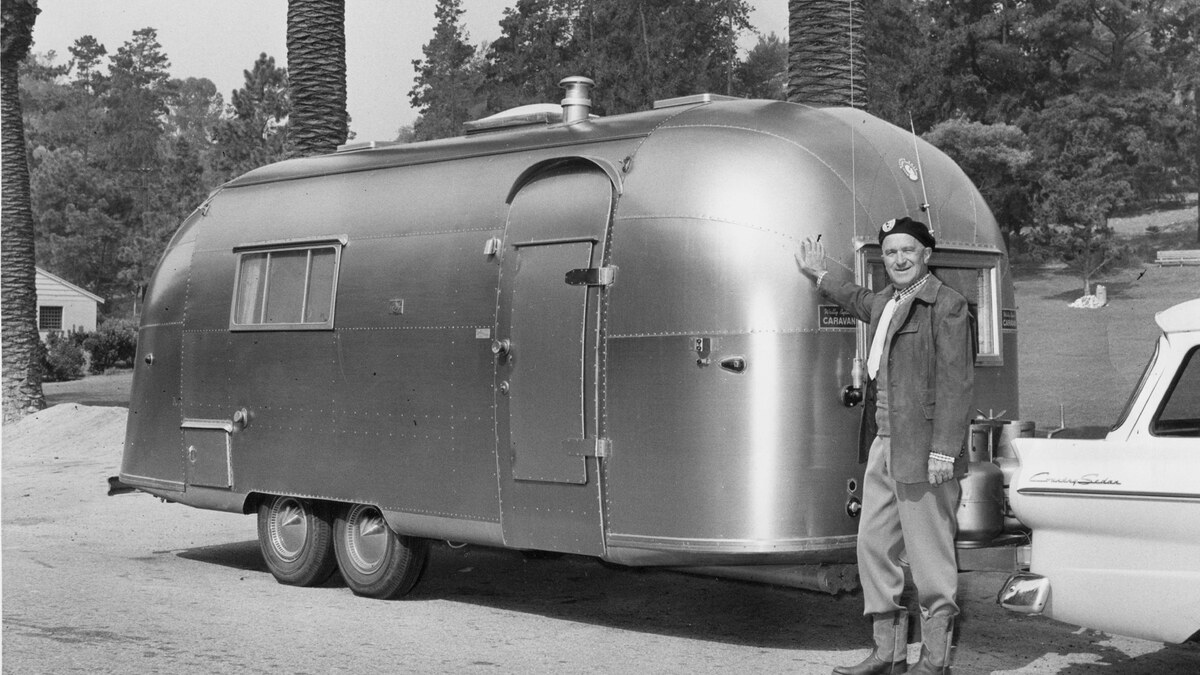 Wally Byam in a dark beret stands beside a polished tandem-axle Airstream caravan trailer.