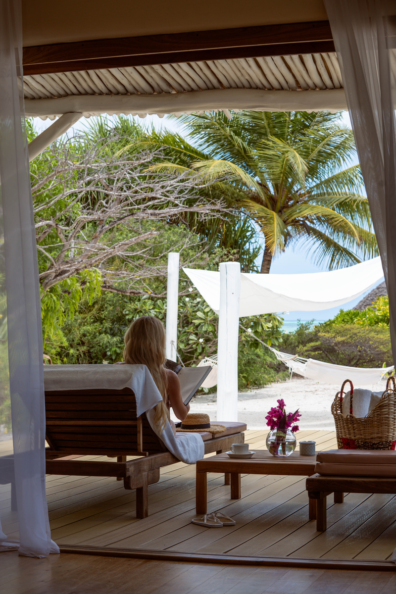 Olga reads on the shaded wooden terrace of her beachfront villa, with a hammock and sea beyond.