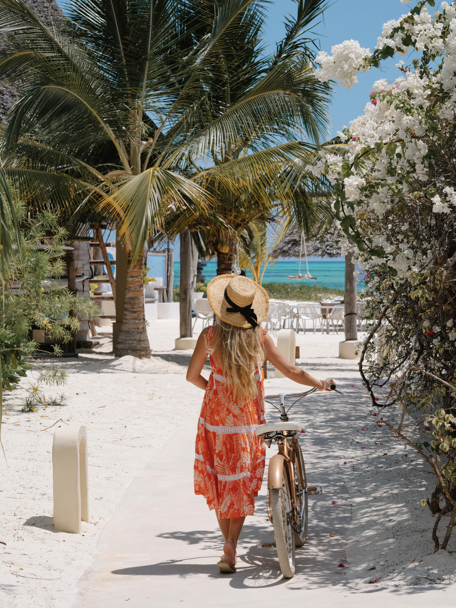 Olga pushes a bicycle along a sandy, flower-lined path towards Paje Beach.