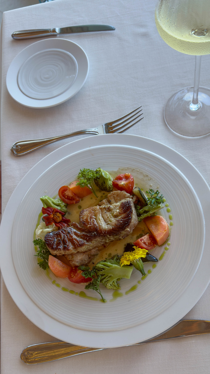 Grilled catch of the day is served with vegetables and edible flowers on a white plate.