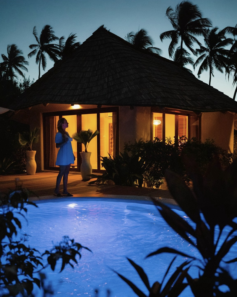 Olga stands beside the blue-lit private pool of her beachfront villa after dark.